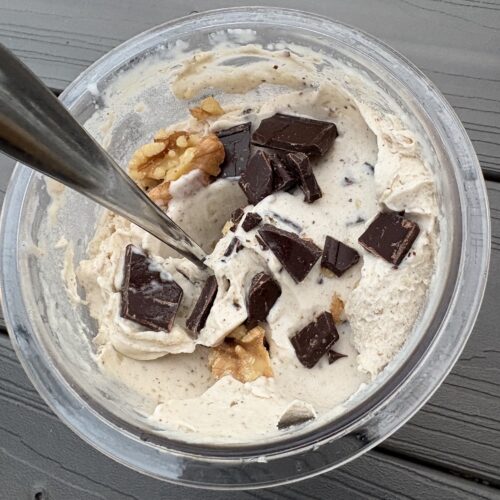 CM Ice Cream | Dana White Nutrition A bowl of vanilla ice cream chunks topped with dark chocolate pieces and cornflakes on a metal dish set on a wooden table, with a spoon visible in the scoop.
