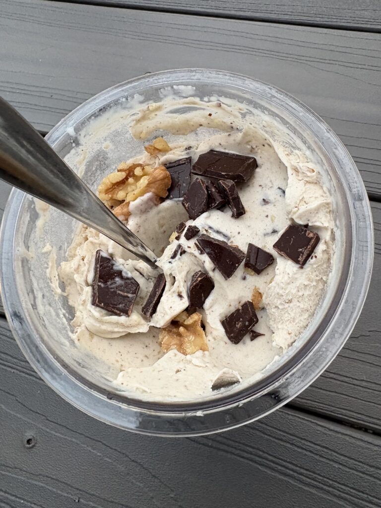 A bowl of vanilla ice cream chunks topped with dark chocolate pieces and cornflakes on a metal dish set on a wooden table, with a spoon visible in the scoop.