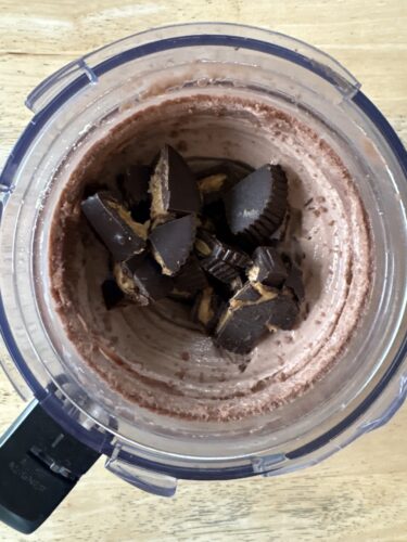 Chopped dark chocolate pieces in a blender jar with a creamy melted chocolate mixture inside, resting on a wooden surface.
