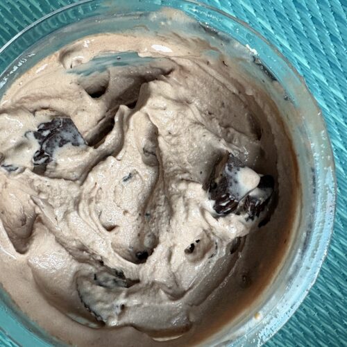 Chocolate mousse with visible cookie pieces in a glass bowl on a teal wicker surface.