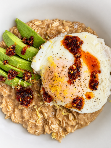 Oatmeal bowl topped with sliced avocado and a fried egg drizzled with chili oil and red pepper flakes.